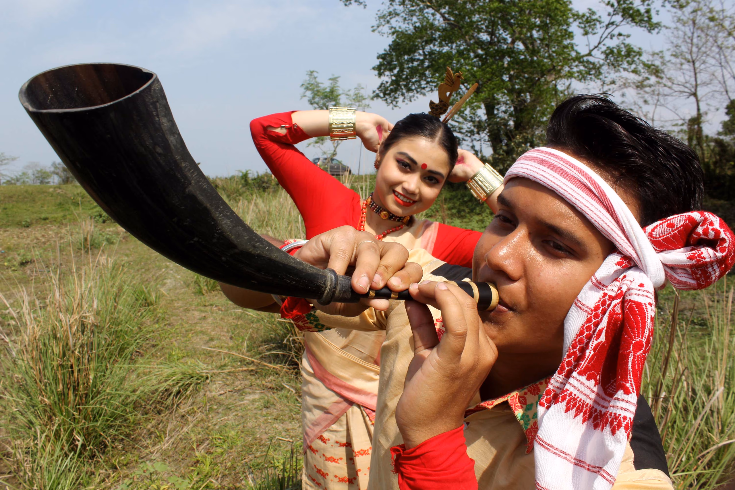 A young man in a Bihu gamosa headband playing the pepa, a buffalo-horn pipe, in close-up, with a woman dancer in red Bihu costume posed behind him in a field