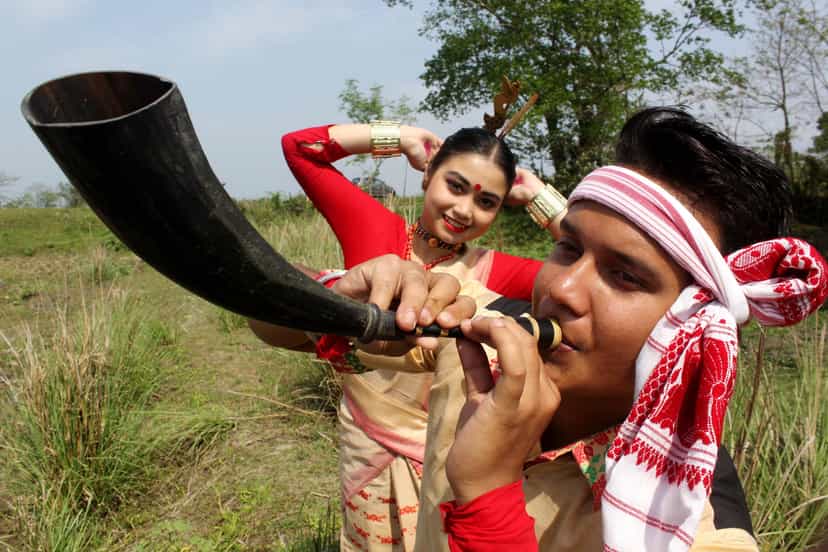 A young man in a Bihu gamosa headband playing the pepa, a buffalo-horn pipe, in close-up, with a woman dancer in red Bihu costume posed behind him in a field