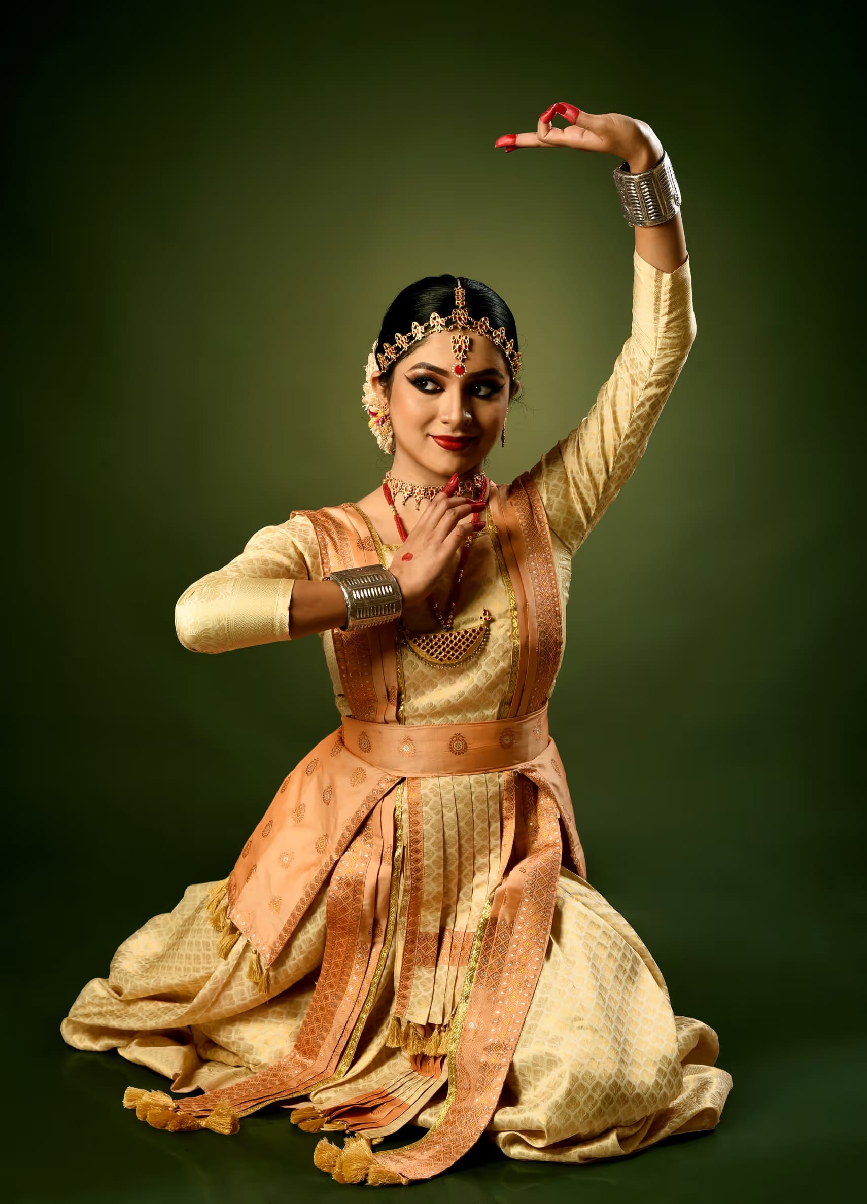 A seated Sattriya dancer in cream pat silk ghuri and blouse, silver gam-kharu cuffs at the wrists, one arm arched overhead in a hand gesture