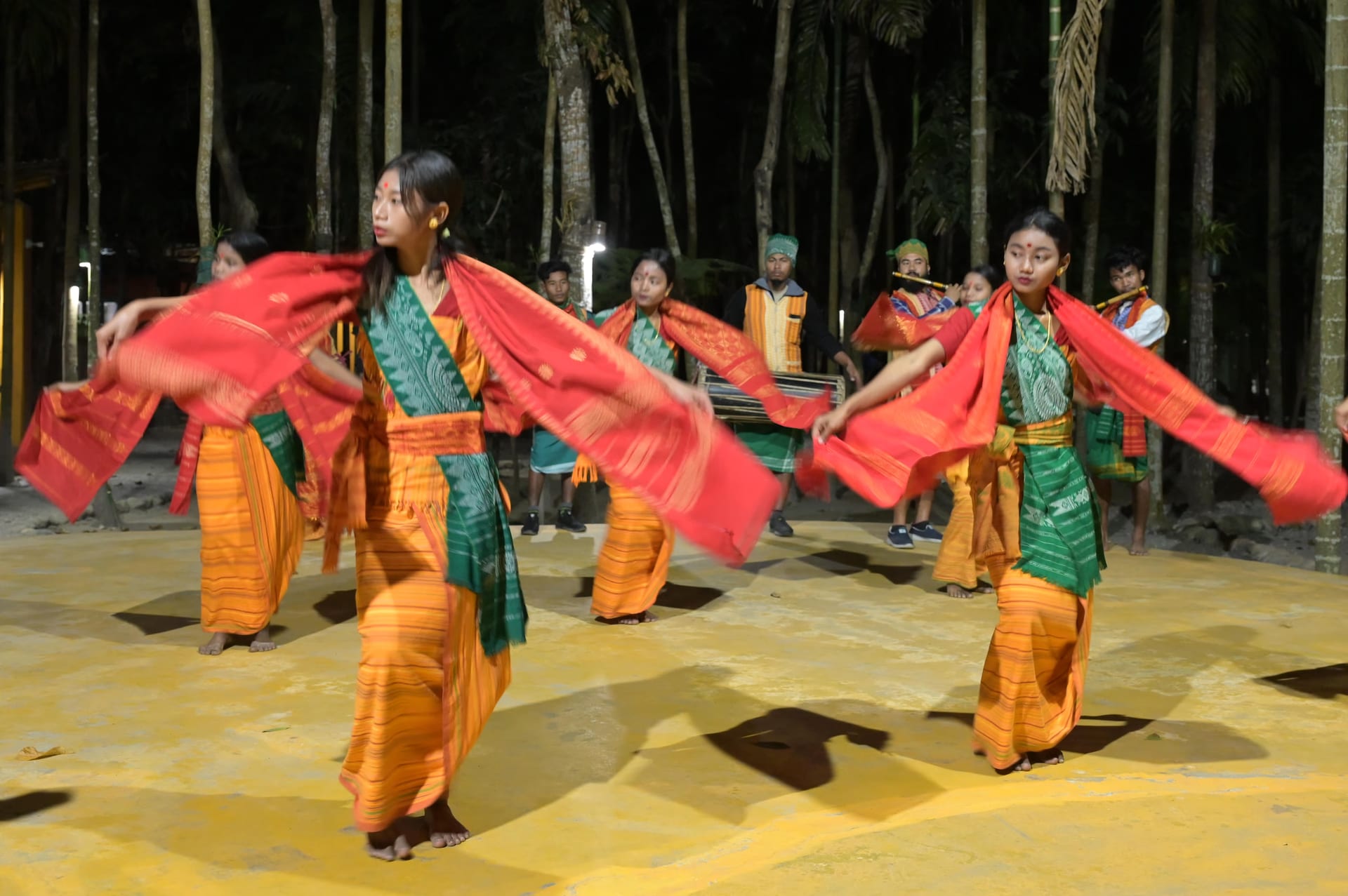 Bodo women dancing the Bagurumba, facing the camera in orange-striped dokhona with green patterned bodices, red aronai scarves stretched wide between their outspread arms
