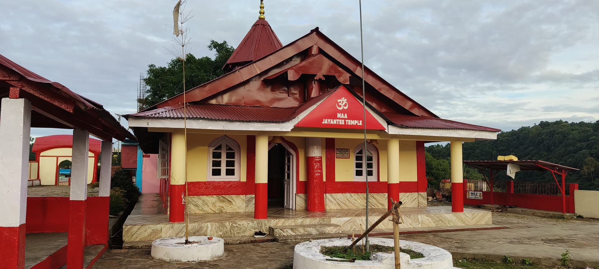 A red-and-cream temple building with a tin roof and a small conical tower, bearing the name Maa Jayantee Temple