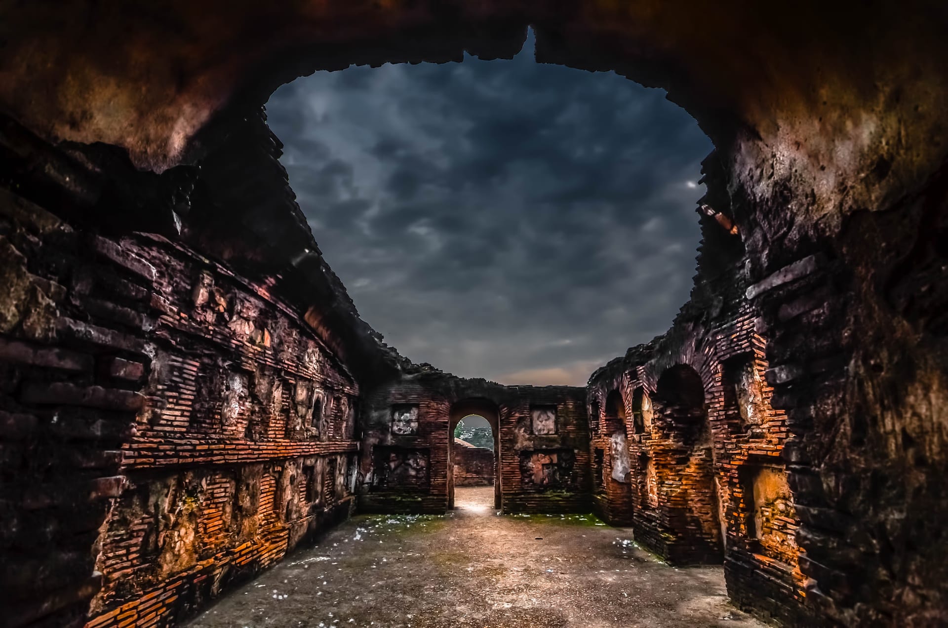 Vaulted brick chamber of the Talatal Ghar opening to a stormy sky through a broken roof, warm-lit golden brick walls and a doorway of light at the far end