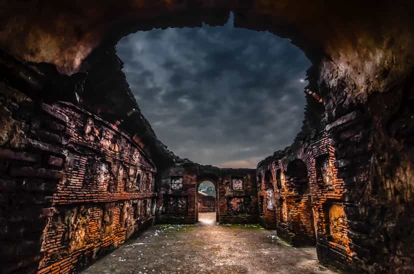 Vaulted brick chamber of the Talatal Ghar opening to a stormy sky through a broken roof, warm-lit golden brick walls and a doorway of light at the far end