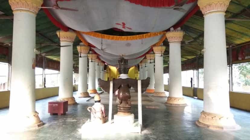 The pillared interior of a Majuli sattra namghar, two rows of whitewashed columns with painted lotus capitals under a draped red-and-white ceiling canopy, a carved wooden throne-pedestal standing in the central aisle on a polished floor