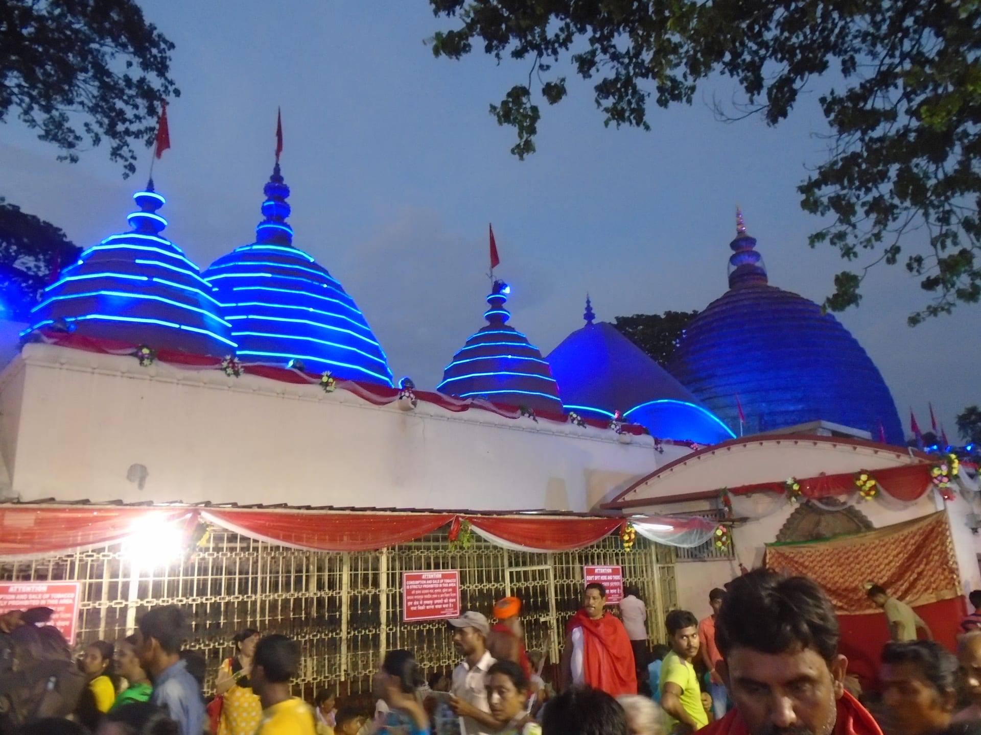 The beehive domes of the Kamakhya temple outlined in blue neon light at dusk, a garlanded gateway below them and festival crowds passing in the foreground during the Ambubachi gathering